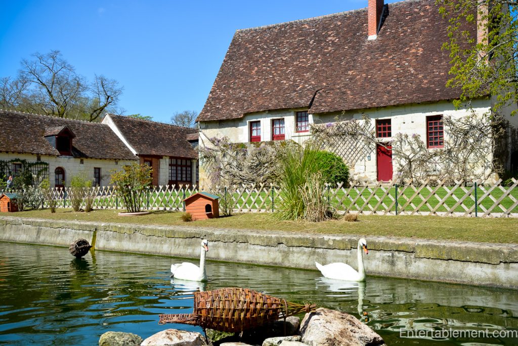 Two white swans glide along a moat in front of historic stone service buildings with red shutters and tiled roofs at Château de Chenonceau.
