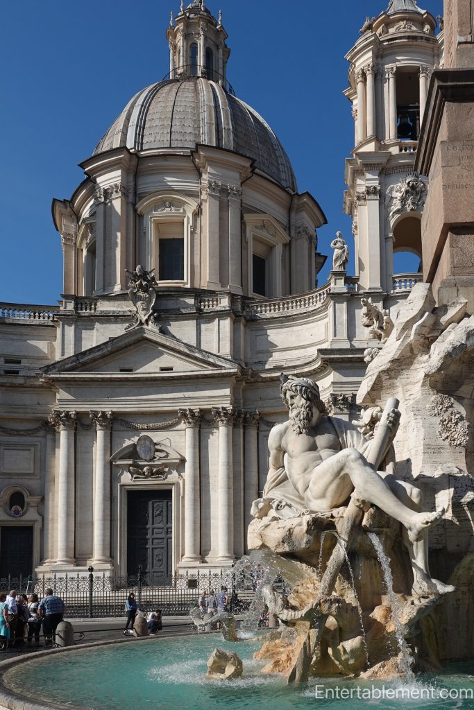 Baroque fountain sculpture of a reclining river god with flowing water in Piazza Navona, Rome, with the domed church of Sant’Agnese in Agone in the background.