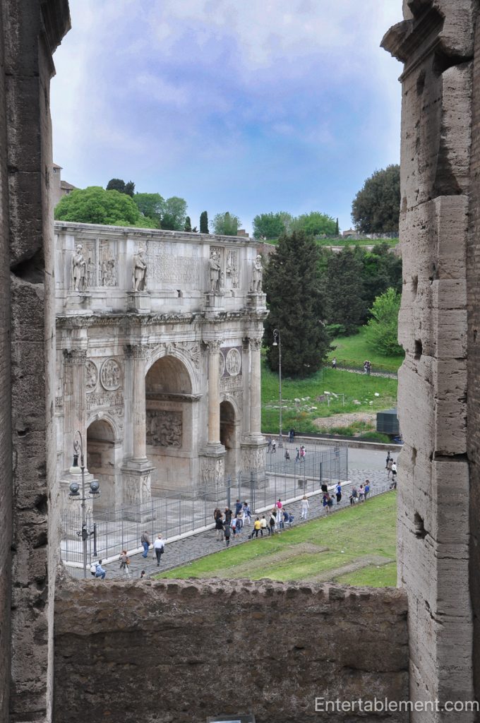 View of the Arch of Constantine seen through a stone opening inside the Colosseum, with carved reliefs, statues, and visitors gathered below.