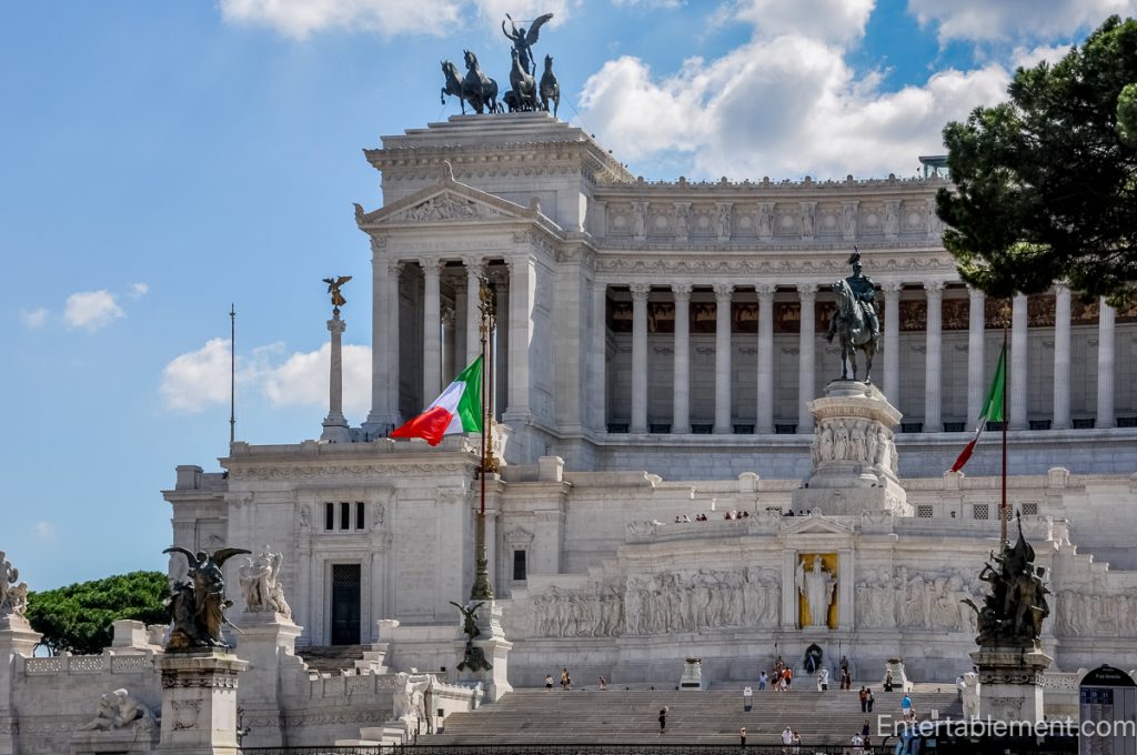 The white marble Vittoriano monument in Rome, with sweeping staircases, sculpted reliefs, Italian flags, and an equestrian statue beneath a blue sky.
