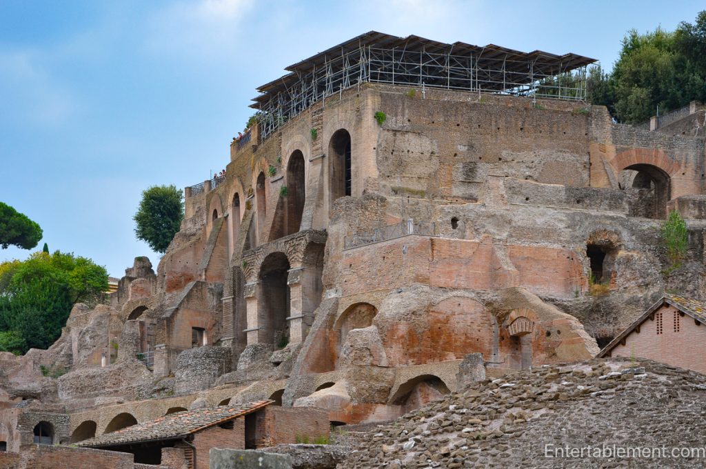 Ancient stone ruins and standing columns in the Roman Forum, surrounded by trees and cypress-lined hills.