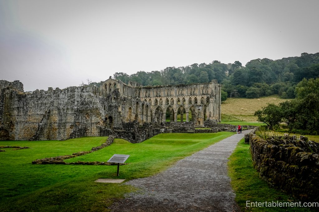 Side view of Rievaulx Abbey ruins set into the valley, with long arcade walls and surrounding hills in the background.